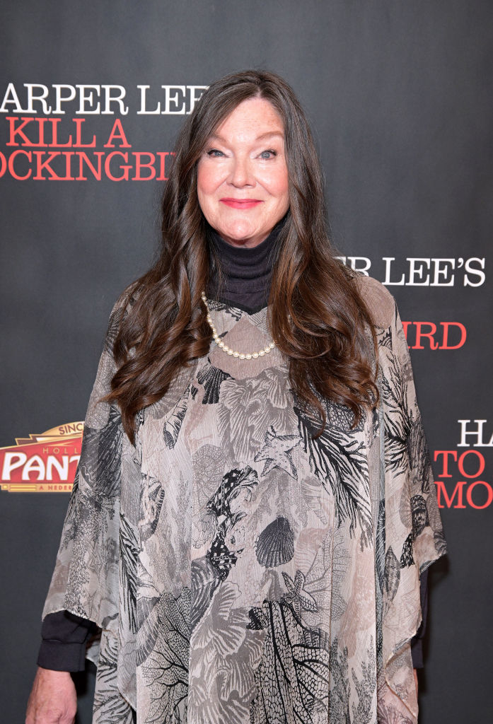 A woman with long hair wearing a patterned poncho stands in front of a backdrop with text "Harper Lee's To Kill a Mockingbird."