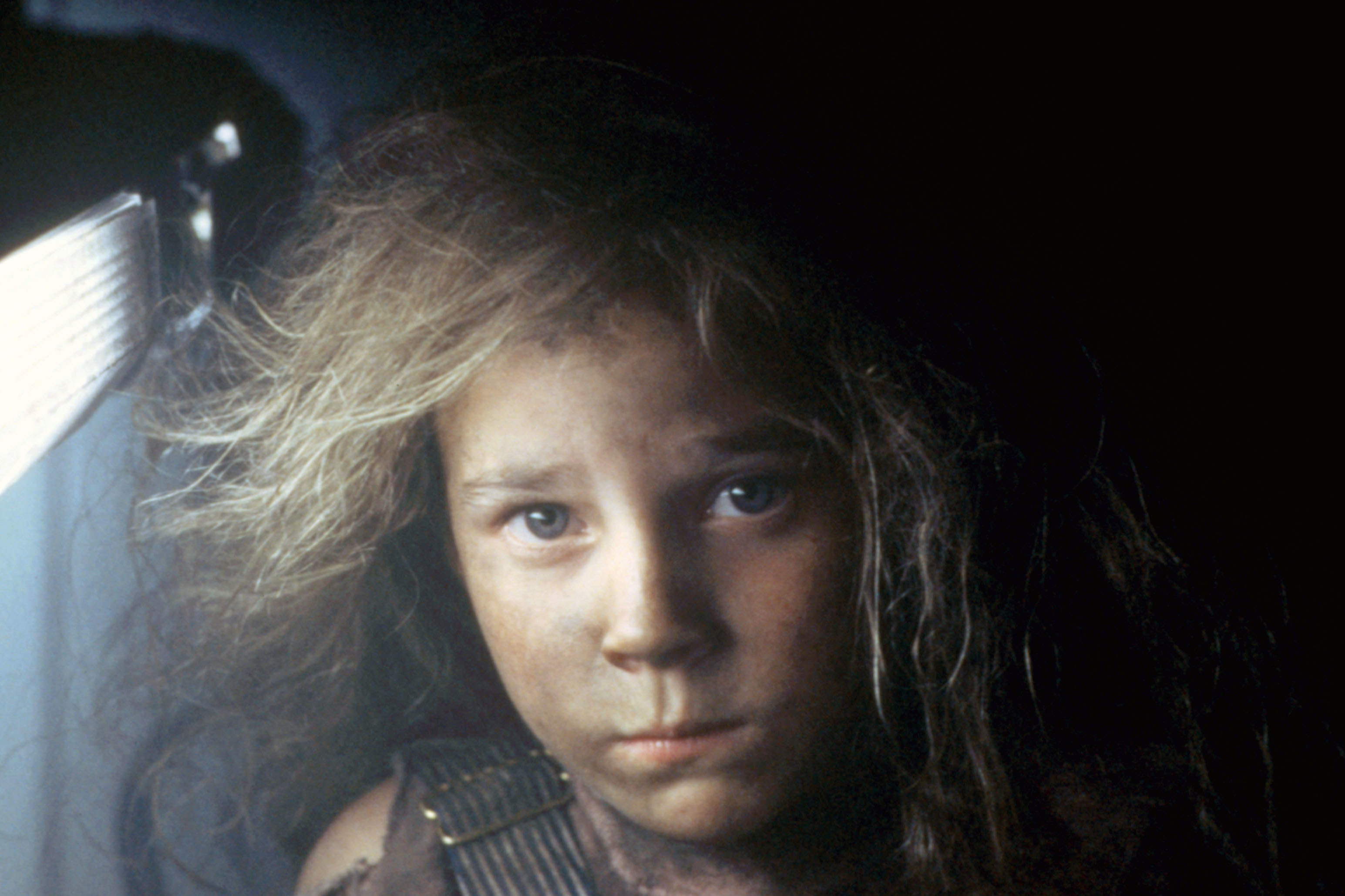 A young girl with disheveled hair and a serious expression, illuminated partially from the left side, stares directly at the camera