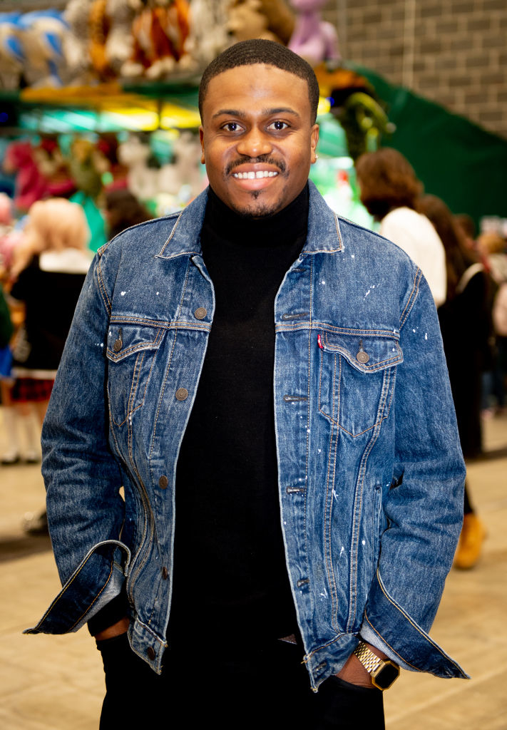 A smiling person stands in a busy indoor setting, wearing a denim jacket over a black turtleneck. Background shows blurred people and colorful items