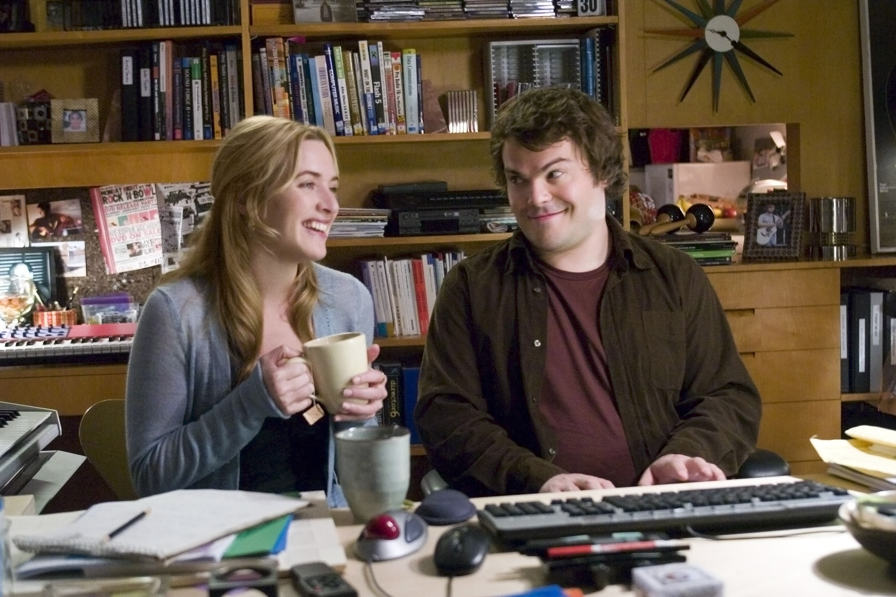 Kate Winslet with a mug and Jack Black with a slight smile, sit at a cluttered desk filled with books, keyboards, and office supplies in a home office setting in "The Holiday"