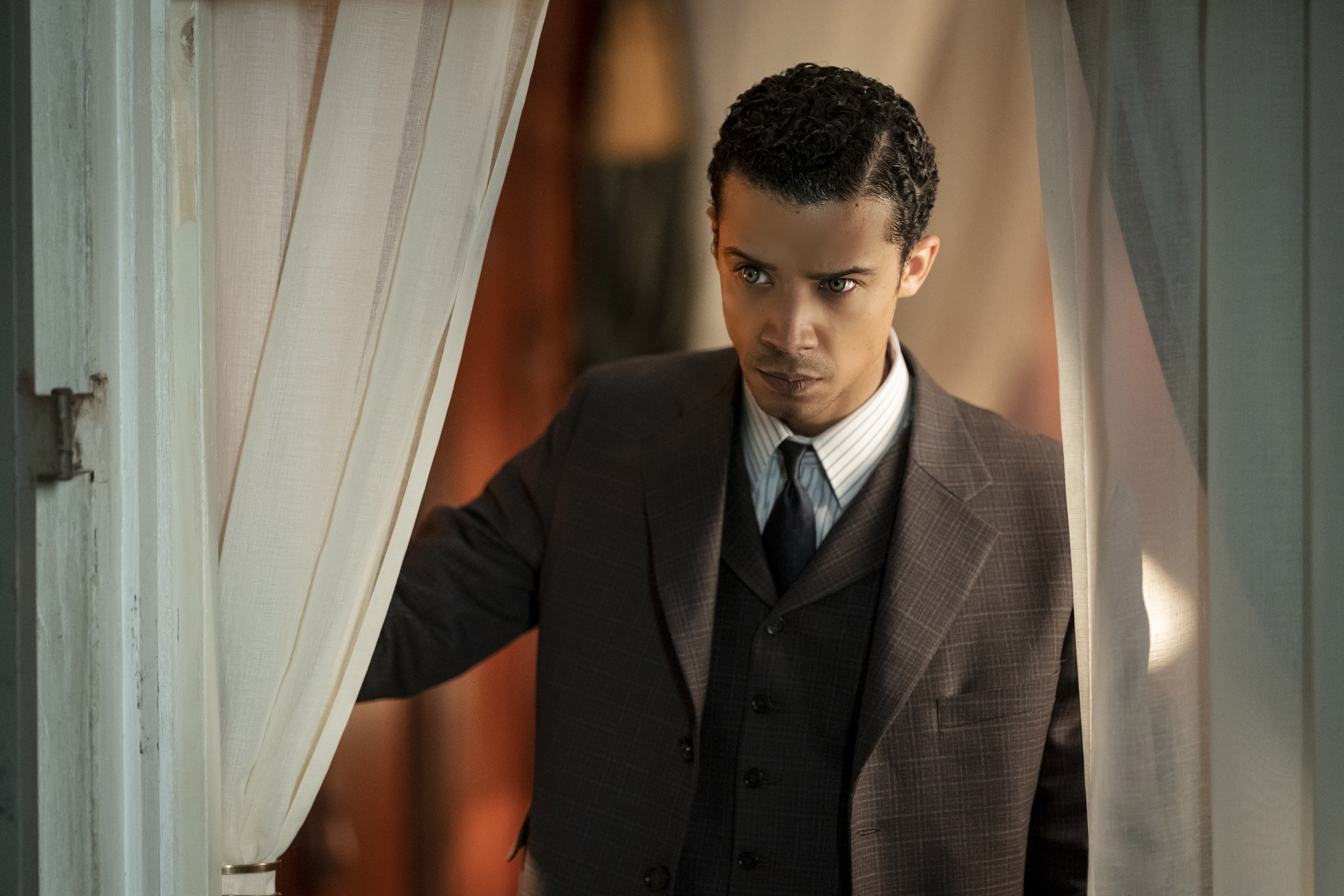 Jacob Anderson wearing a classic brown suit and tie, stands in a doorway with white curtains in a scene from &quot;Interview with the Vampire&quot;