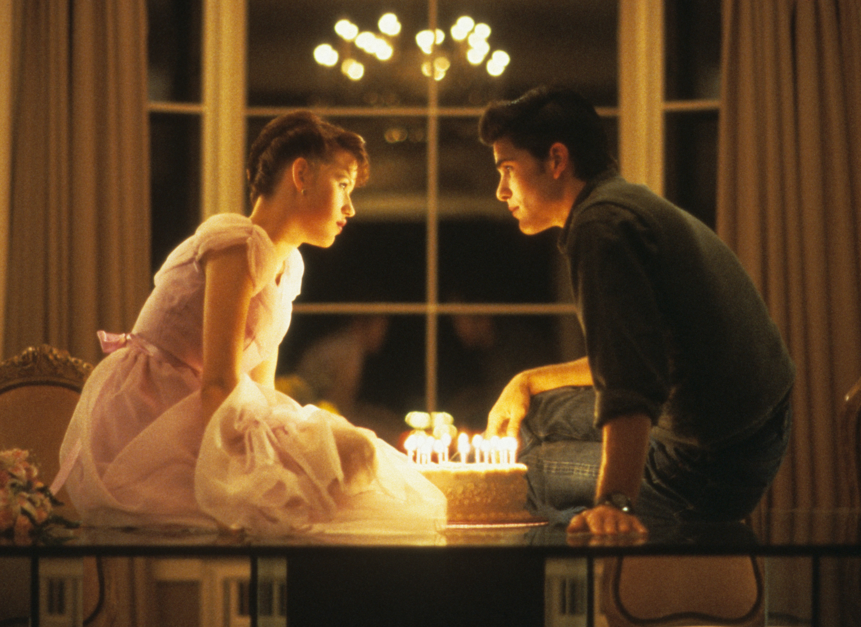 Molly Ringwald and Michael Schoeffling sit on a table with a lit birthday cake between them in a scene from "Sixteen Candles"
