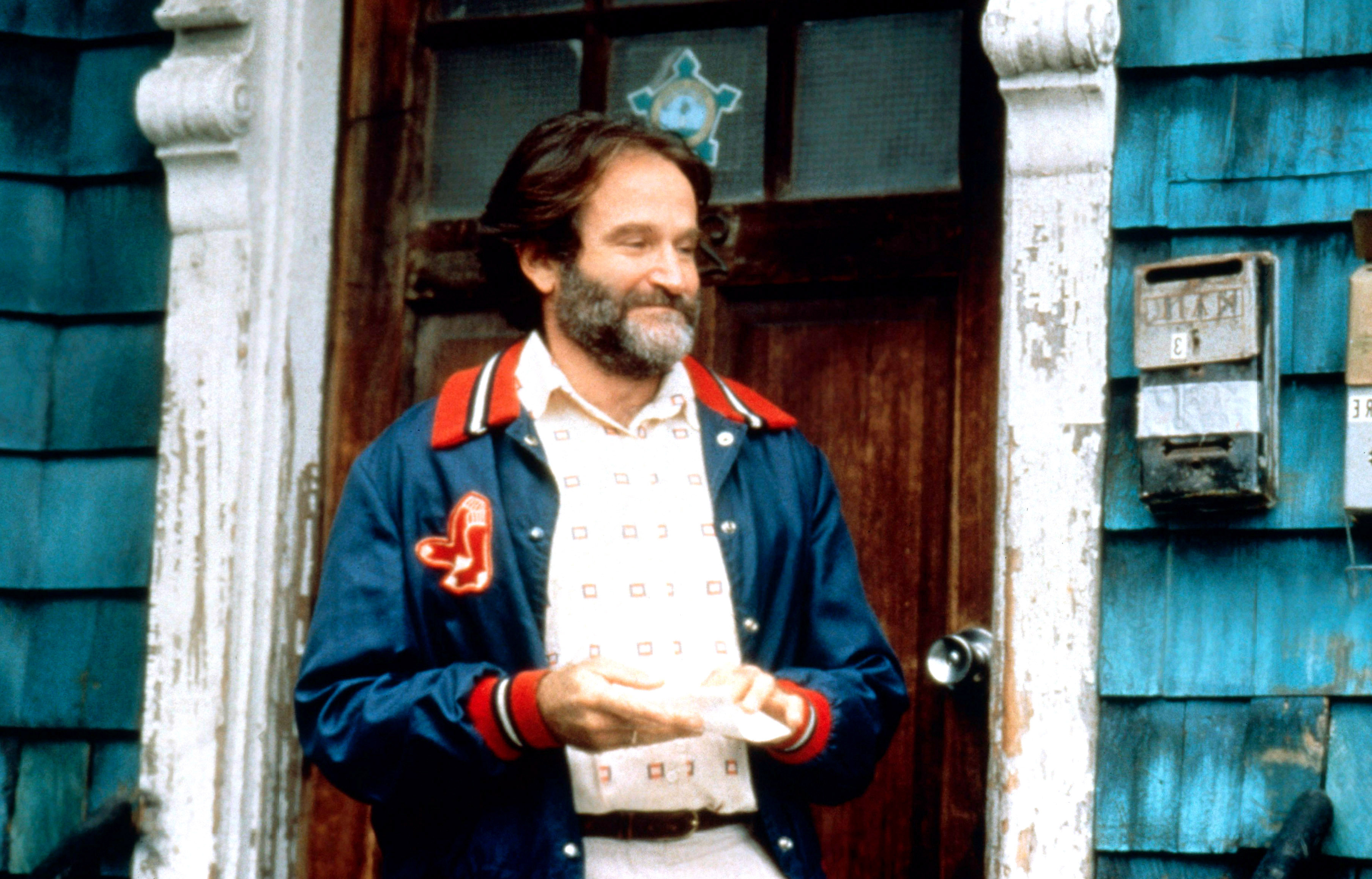 Robin Williams stands in front of a weathered building, wearing a patterned shirt under a jacket with a red collar and cuffs, smiling warmly