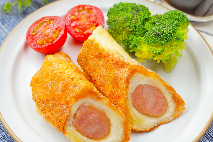 お皿に盛られたソーセージをパンで包んで揚げた料理、ブロッコリーとトマトが添えられている。