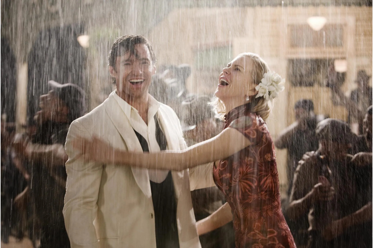 Hugh Jackman in a white suit and Nicole Kidman in an elegant dress, both laughing and dancing in the rain amidst a bustling street scene