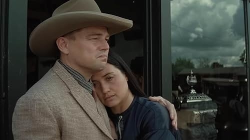 Leonardo DiCaprio, in a suit and cowboy hat, embraces Lily Gladstone, in casual attire, as they stand next to a glass door