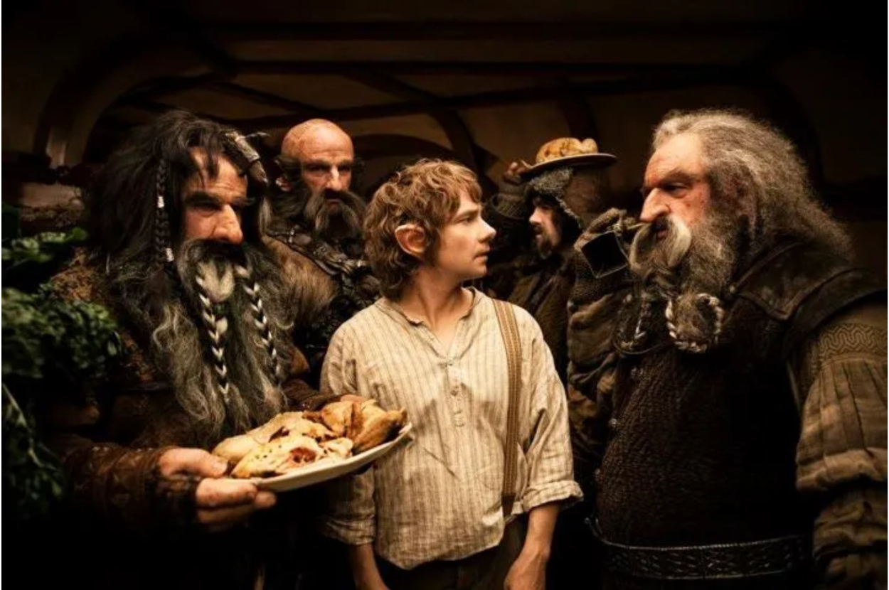Dwarves Dwalin, Bofur, and Thorin Oakenshield stand with Bilbo Baggins in a scene from "The Hobbit." They are conversing, and one dwarf holds a plate of food