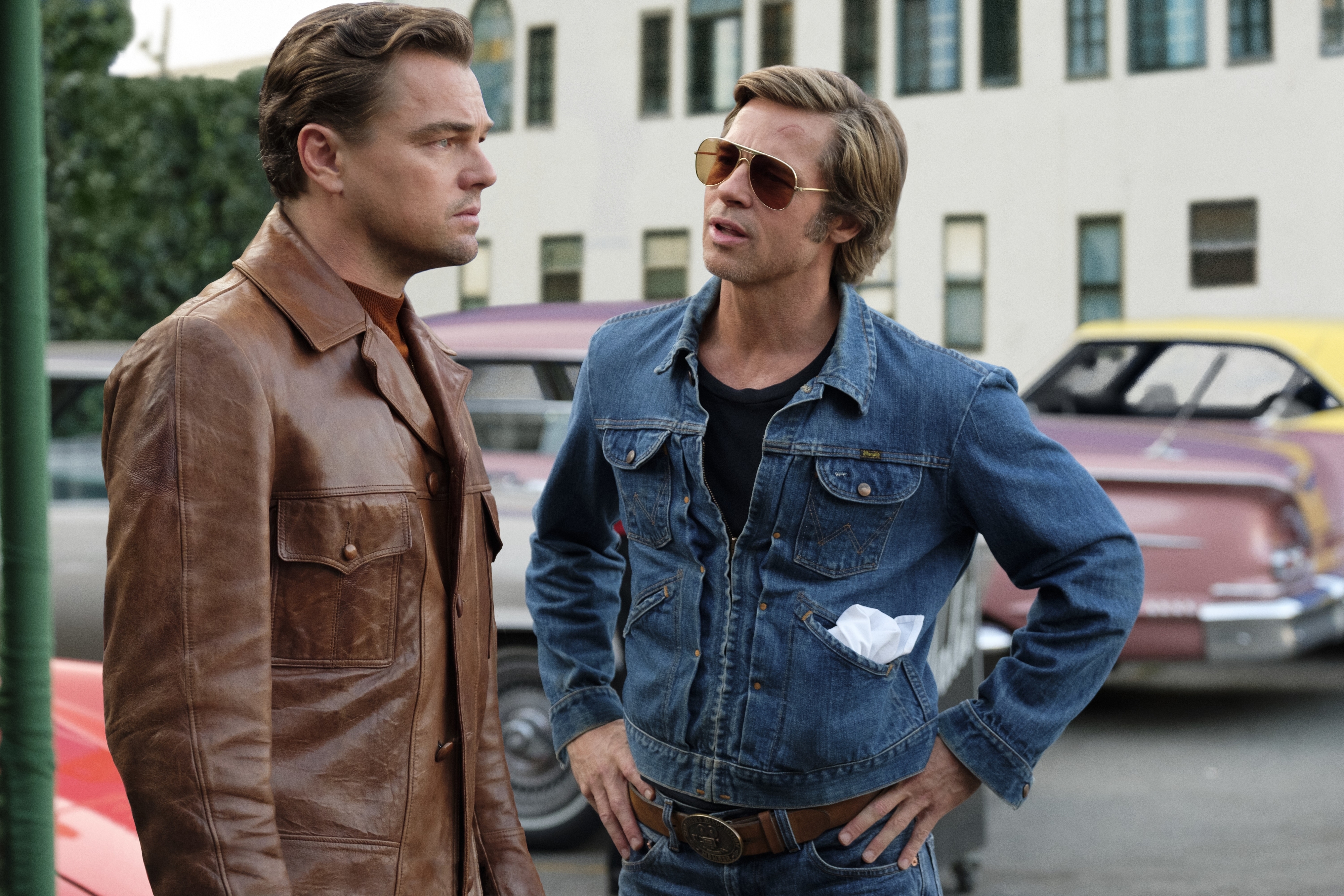 Leonardo DiCaprio in a brown leather jacket and Brad Pitt in a denim jacket, talking in front of vintage cars on a movie set