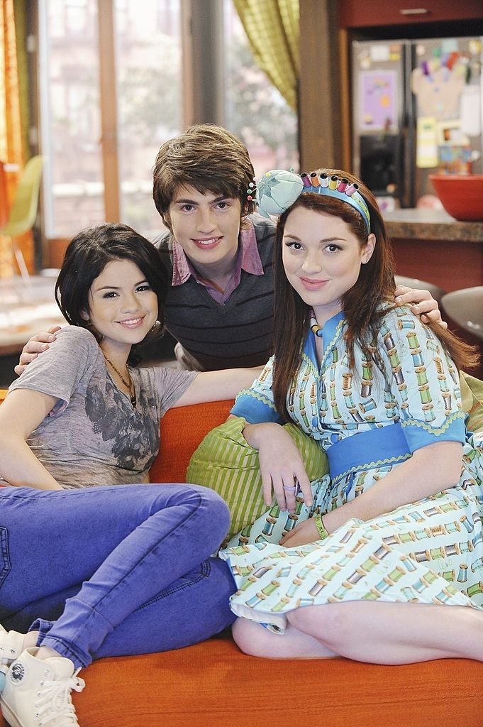 Selena Gomez, David Henrie, and Jennifer Stone smile while sitting on a couch. Selena wears a casual shirt and jeans, David wears a sweater, and Jennifer wears a dress