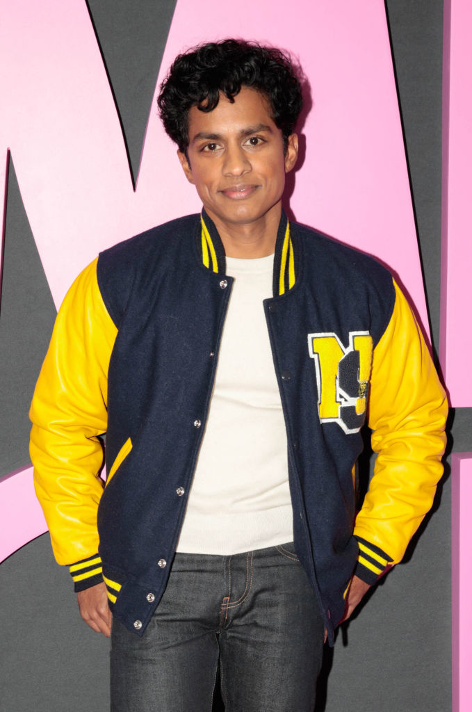 Naveen Andrews wearing a letterman jacket with yellow sleeves, a white shirt, and dark jeans, poses in front of a pink and black background