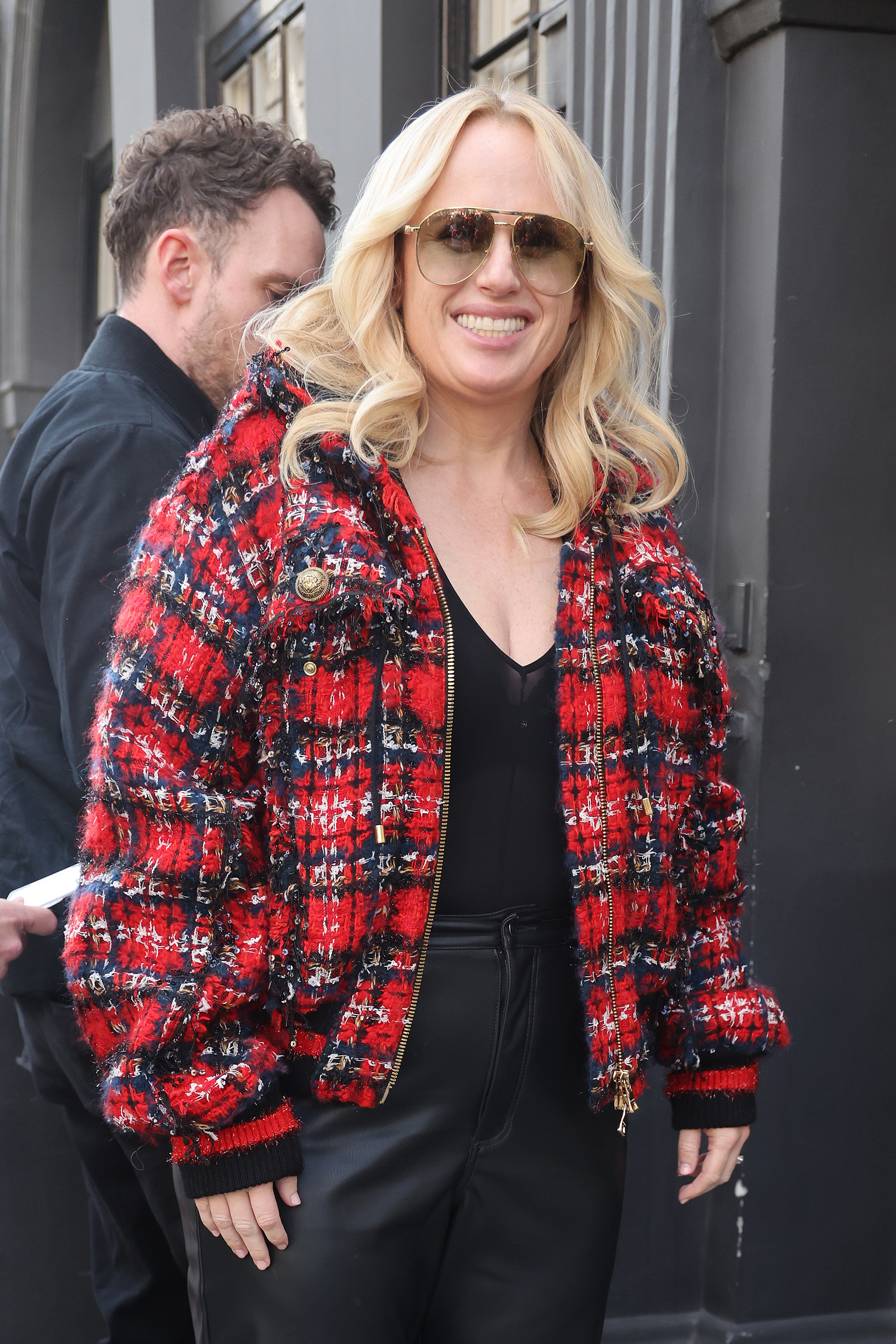 Rebel Wilson smiles wearing a red plaid jacket with black leather pants and sunglasses, accompanied by an unidentified man in a dark outfit