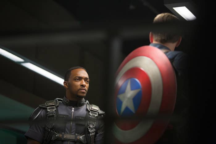 Anthony Mackie in a tactical suit faces Chris Evans, who wears Captain America's shield on his back. They appear to be in a dimly lit, serious setting
