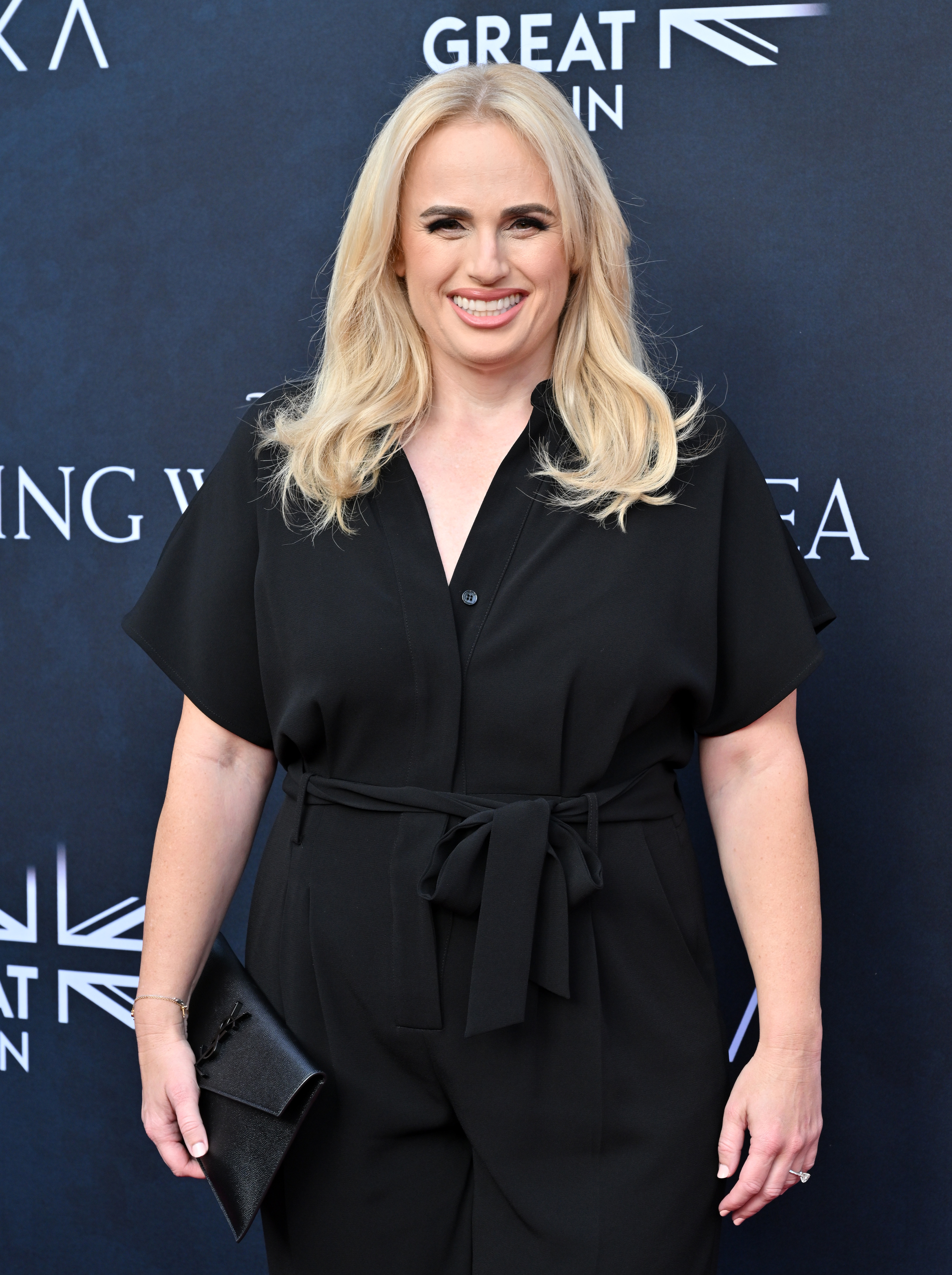 Rebel Wilson smiling on the red carpet, wearing a black jumpsuit with short sleeves and a tied waist, holding a black clutch