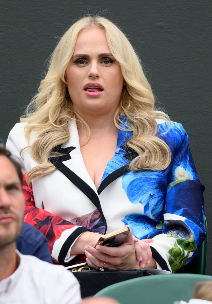 Rebel Wilson watching a tennis match, seated and holding a phone. She is wearing a floral blazer with bold patterns