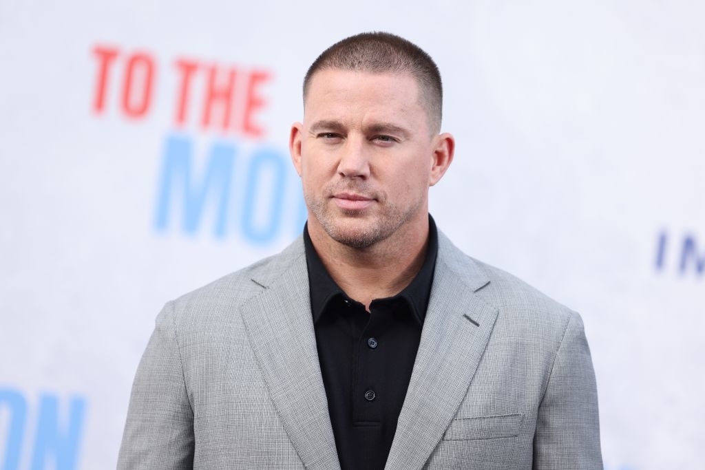 Channing Tatum at an event, wearing a light gray suit jacket over a black shirt, posing in front of a backdrop with partial text visible