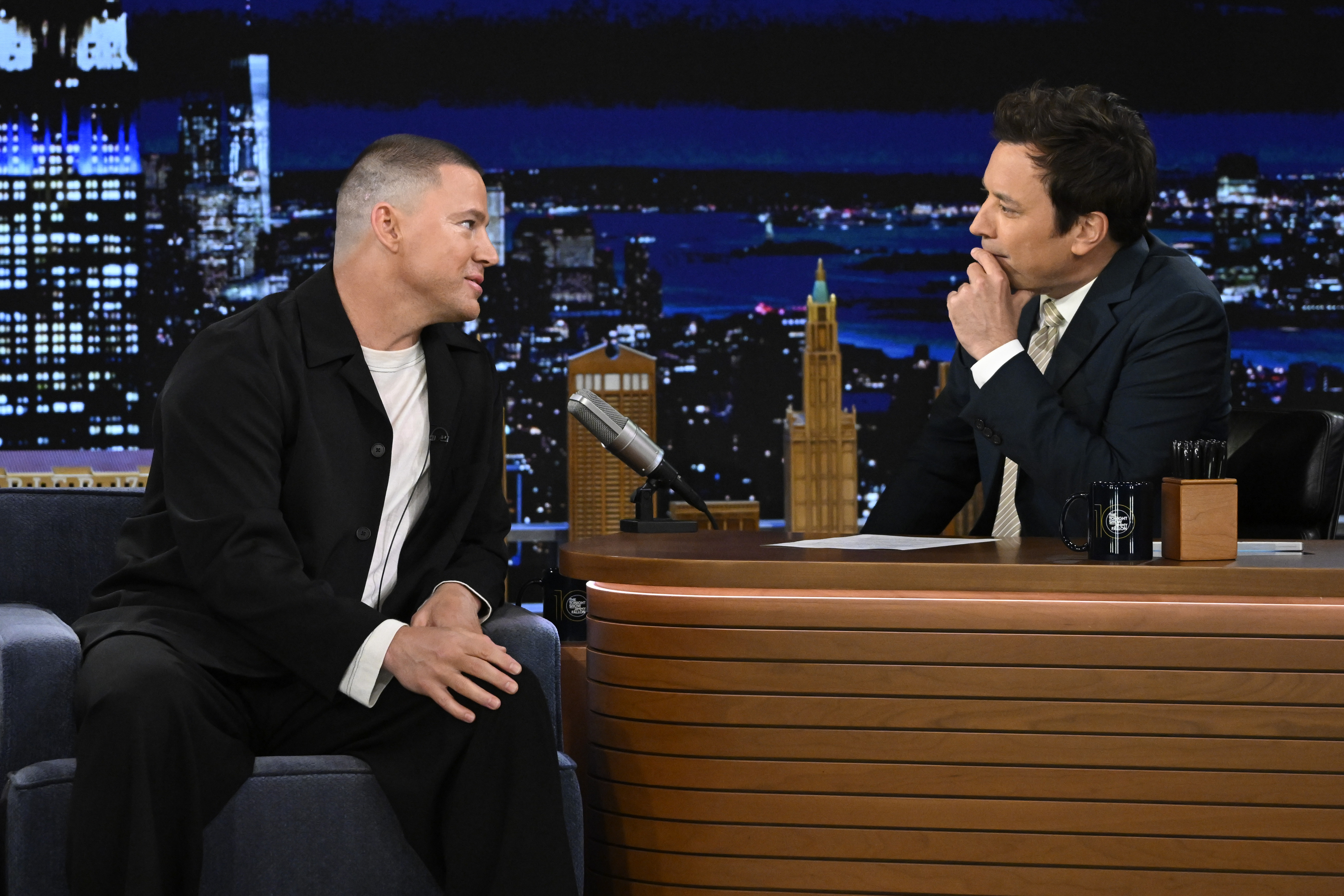 Channing Tatum in a casual suit being interviewed by Jimmy Fallon in a formal suit on a late-night talk show set resembling a cityscape backdrop