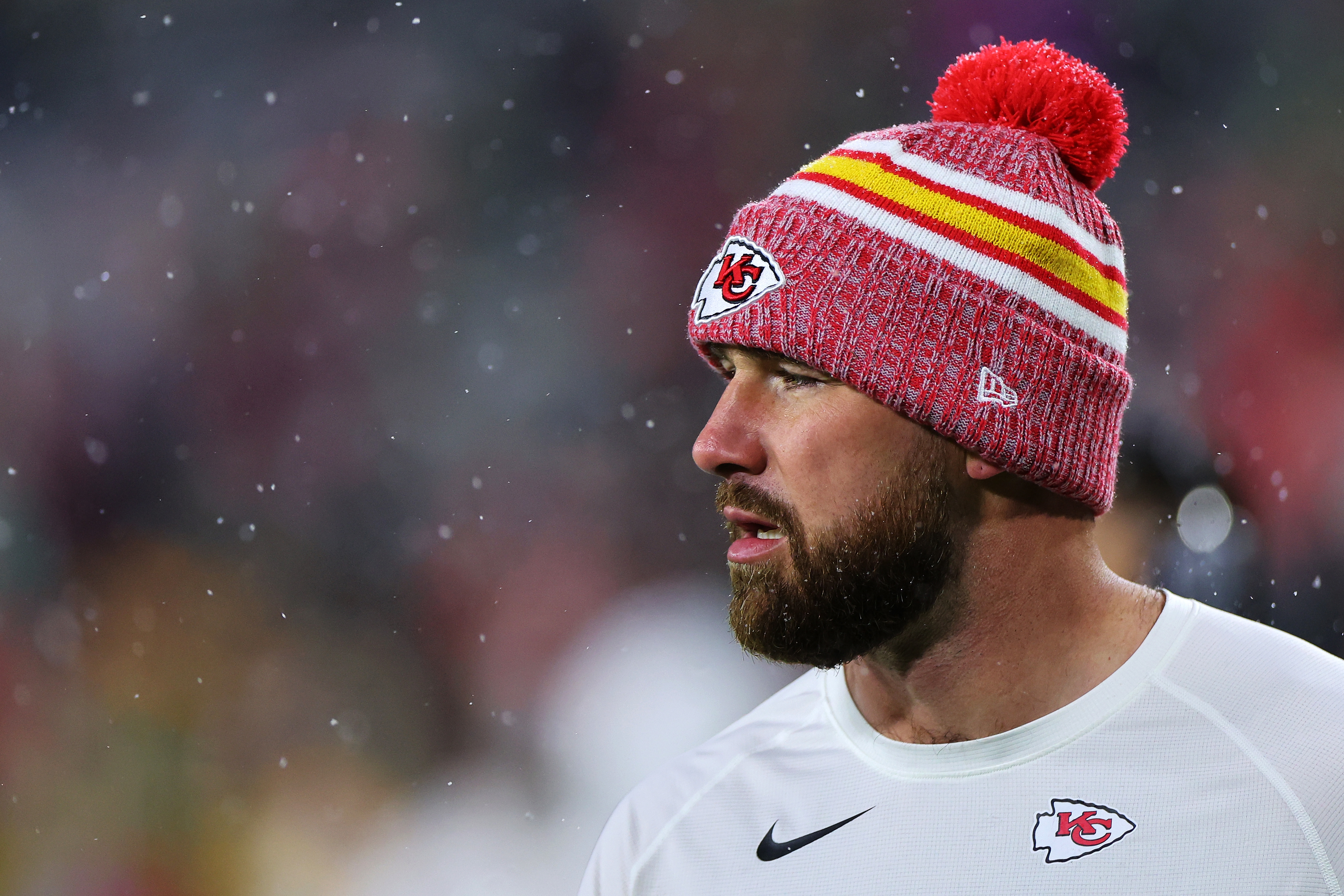 Travis Kelce, wearing a Kansas City Chiefs knit hat and white sports shirt, looking to the right. Snowflakes are falling around him