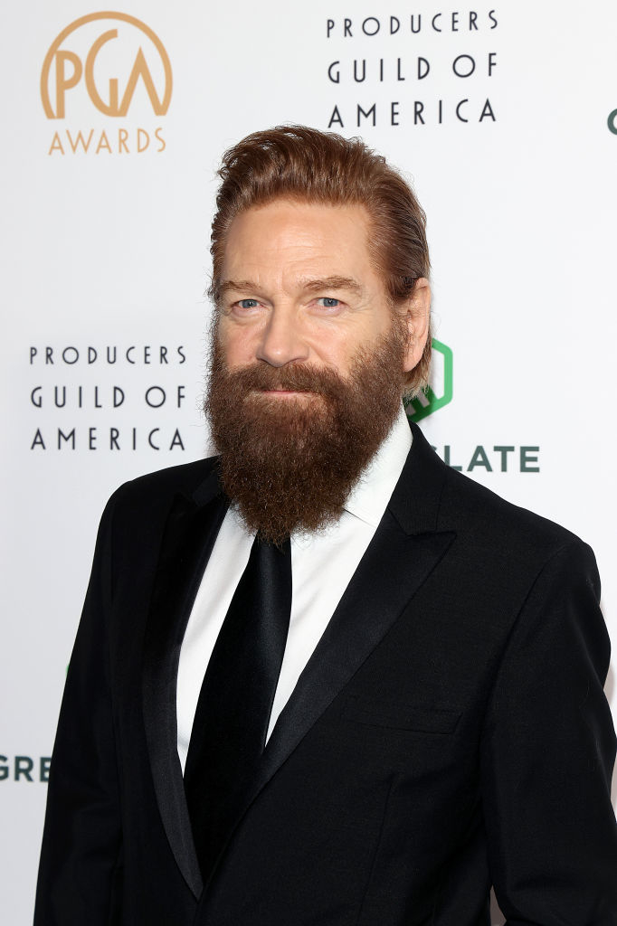 A man with a thick beard and styled hair is wearing a black suit with a white shirt and black tie, posing at the Producers Guild of America Awards