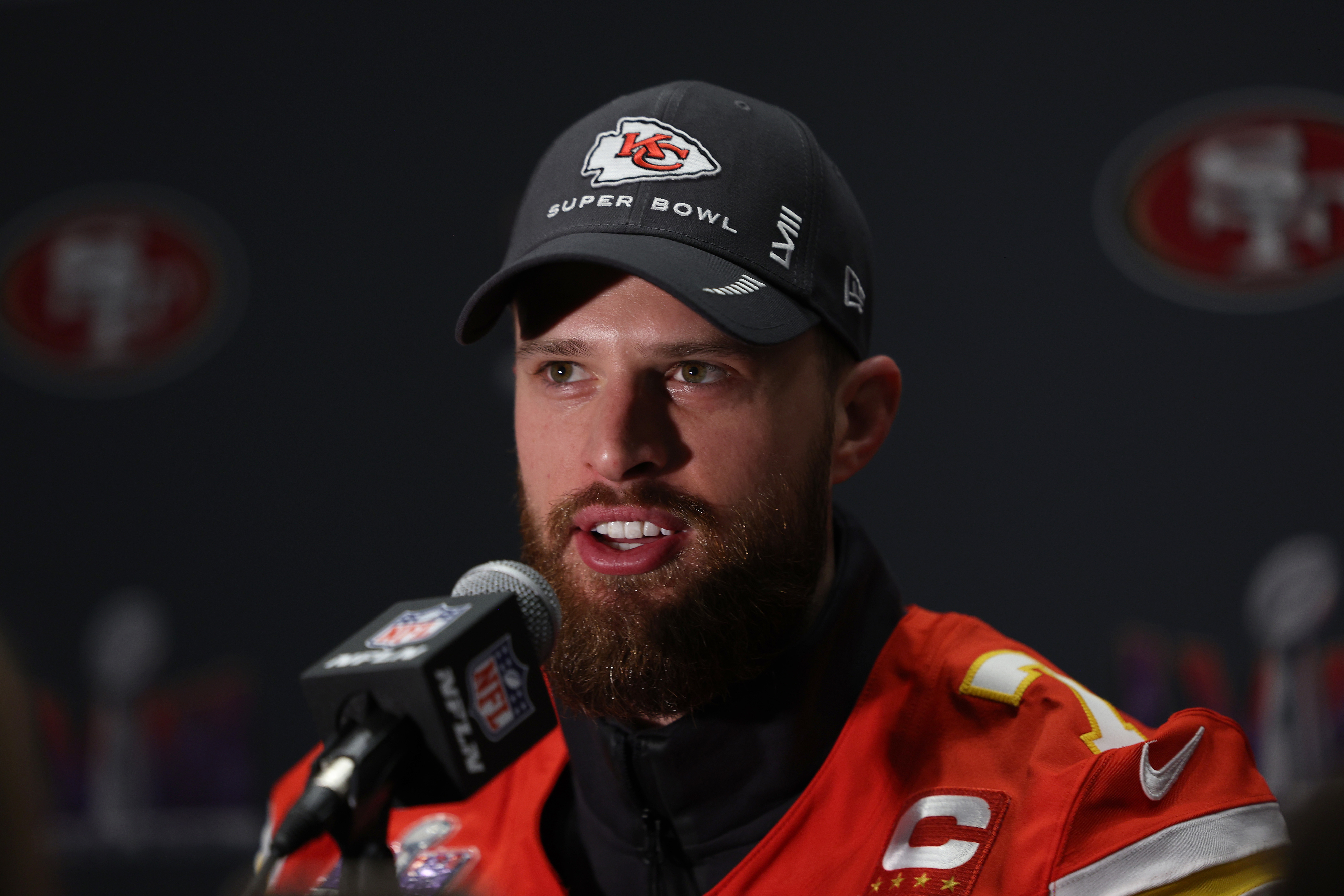 Harrison Butker wearing a Kansas City Chiefs jacket and Super Bowl LVII hat speaks into a microphone at a press event