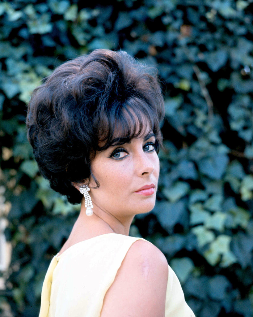 Elizabeth Taylor with a styled updo and pearl drop earrings, poses outdoors with a leafy backdrop, looking over her shoulder
