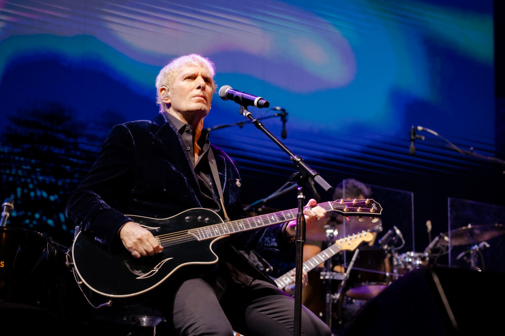 Michael Bolton performs on stage, playing a guitar, with a focused expression