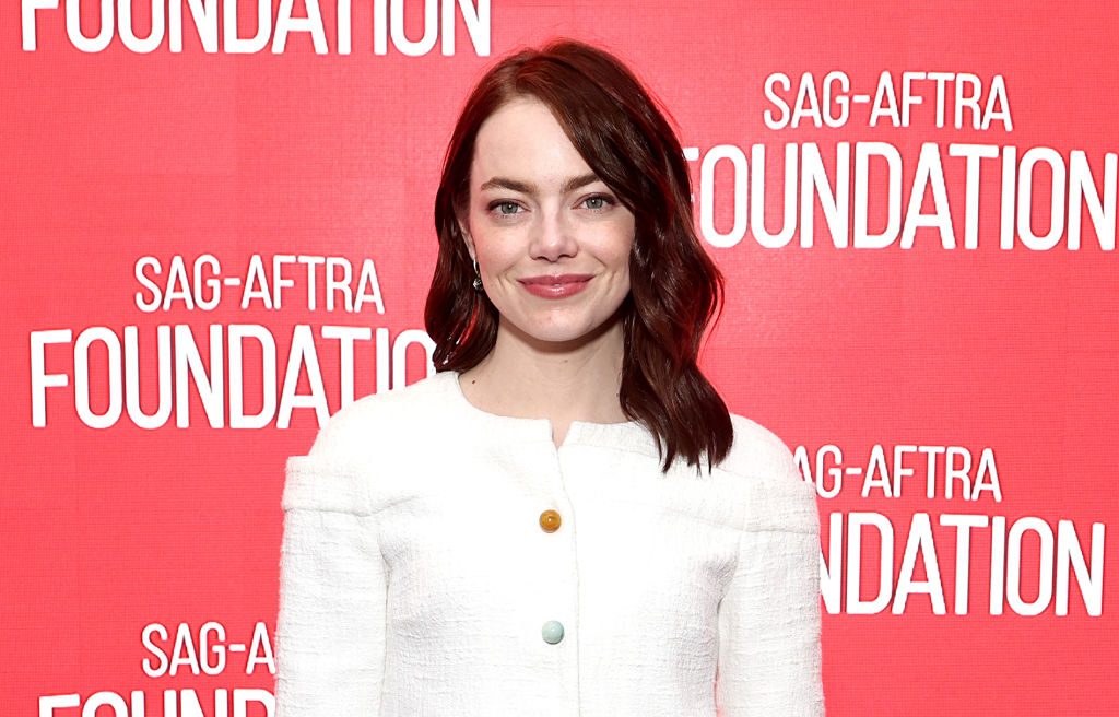Emma Stone standing in front of a SAG-AFTRA Foundation backdrop, wearing a stylish white outfit and smiling at the camera