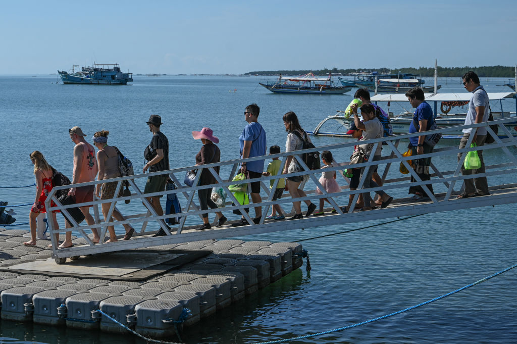 People walking along a dock, boarding a boat in a coastal area. Water, boats, and distant shoreline are visible in the background