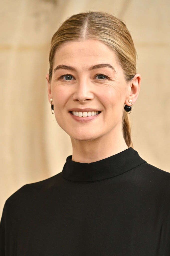 Rosamund Pike smiling, wearing a simple, elegant black high-neck dress with small earrings