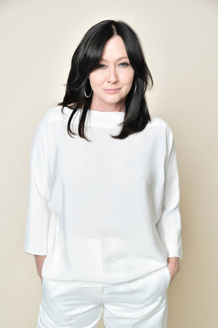 Shannen Doherty in a white, loose-fitting top and matching pants, posing with hands in pockets against a neutral background