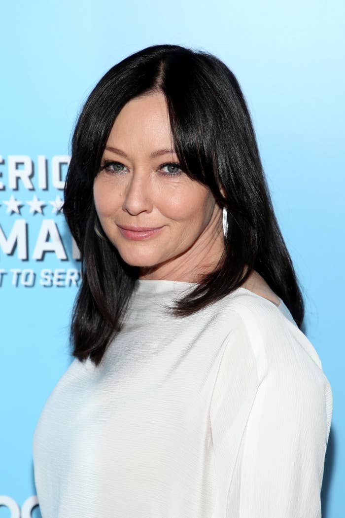 Shannen Doherty smiling, wearing a simple, elegant long-sleeved white dress in front of a blue background