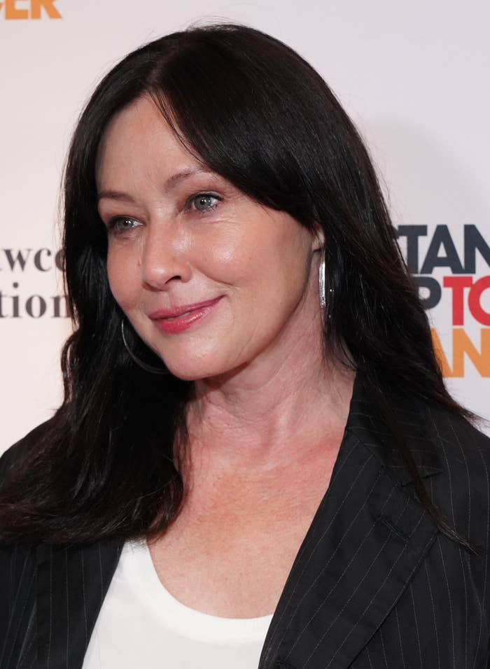 Shannen Doherty at a Stand Up To Cancer event, wearing a black pinstripe blazer and hoop earrings