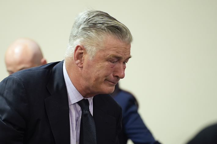 Alec Baldwin is seen in a courtroom, looking emotional while wearing a dark suit, light shirt, and dark tie. A bald person sits in the background