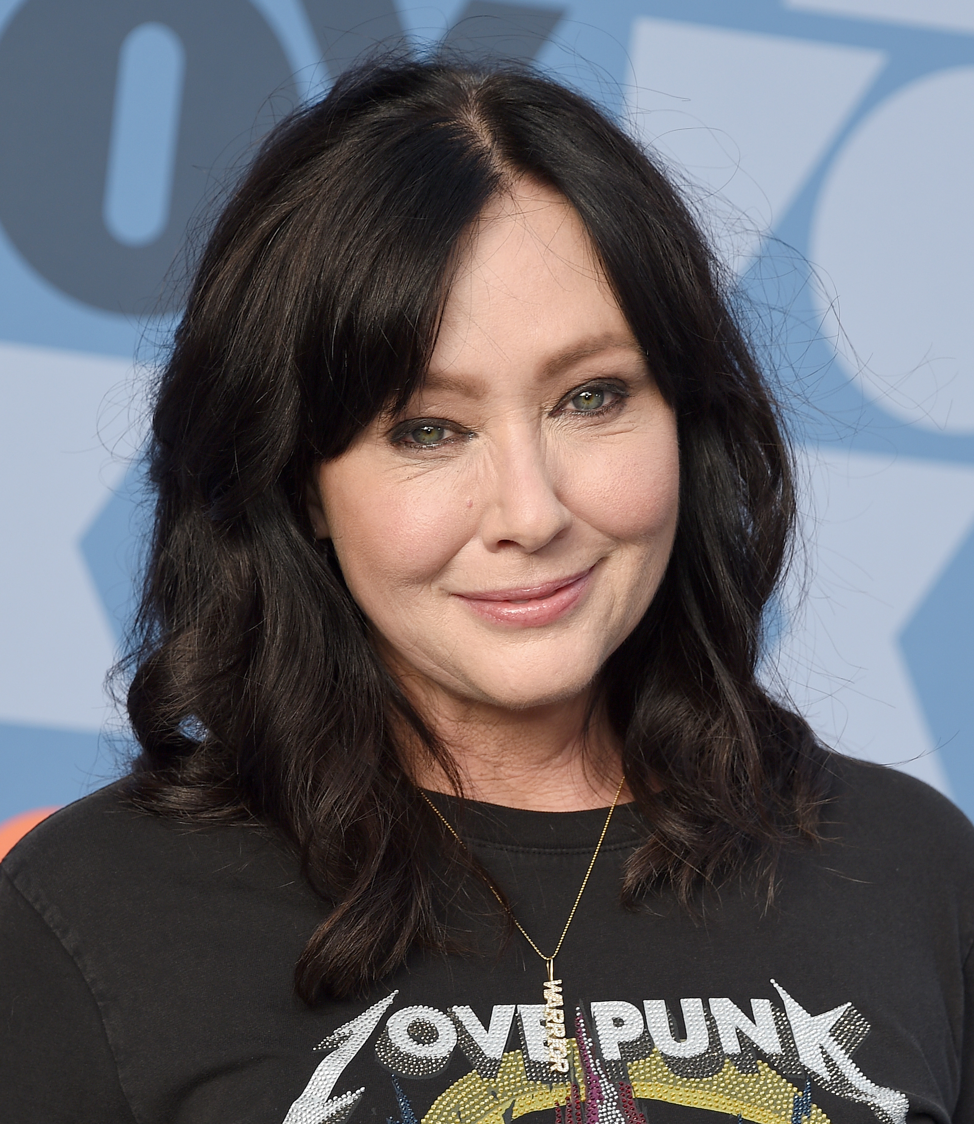 Shannen Doherty smiling at an event, wearing a "LOVE PUNK" graphic black t-shirt, with wavy hair styled down