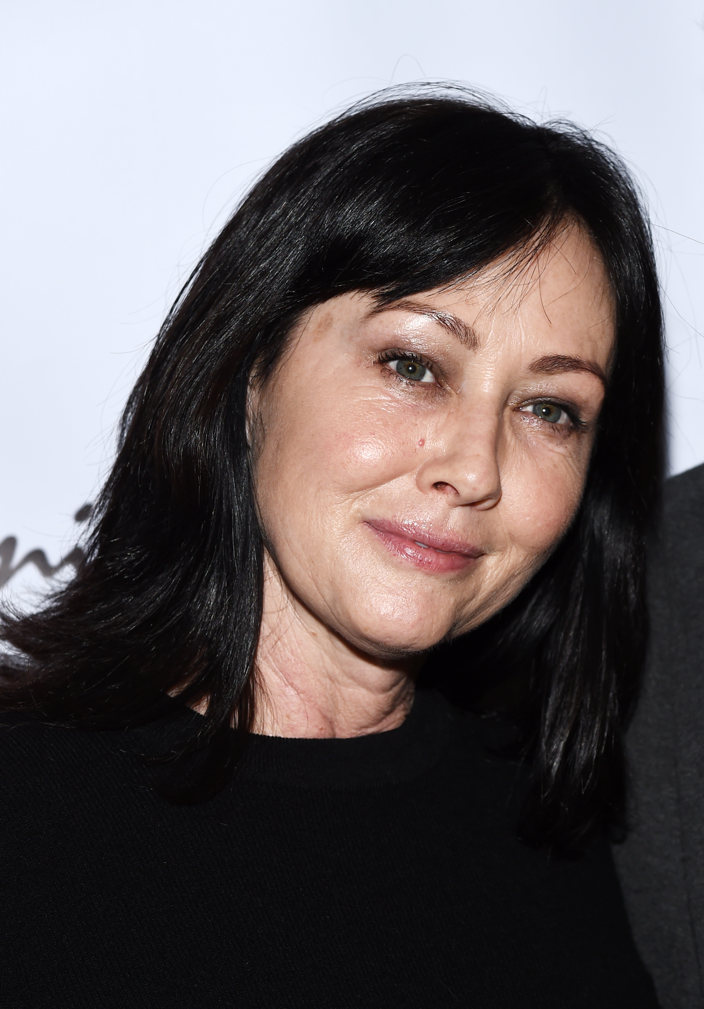 Shannen Doherty smiles softly at the camera, with her dark hair framing her face. She is wearing a black top and standing against a plain background