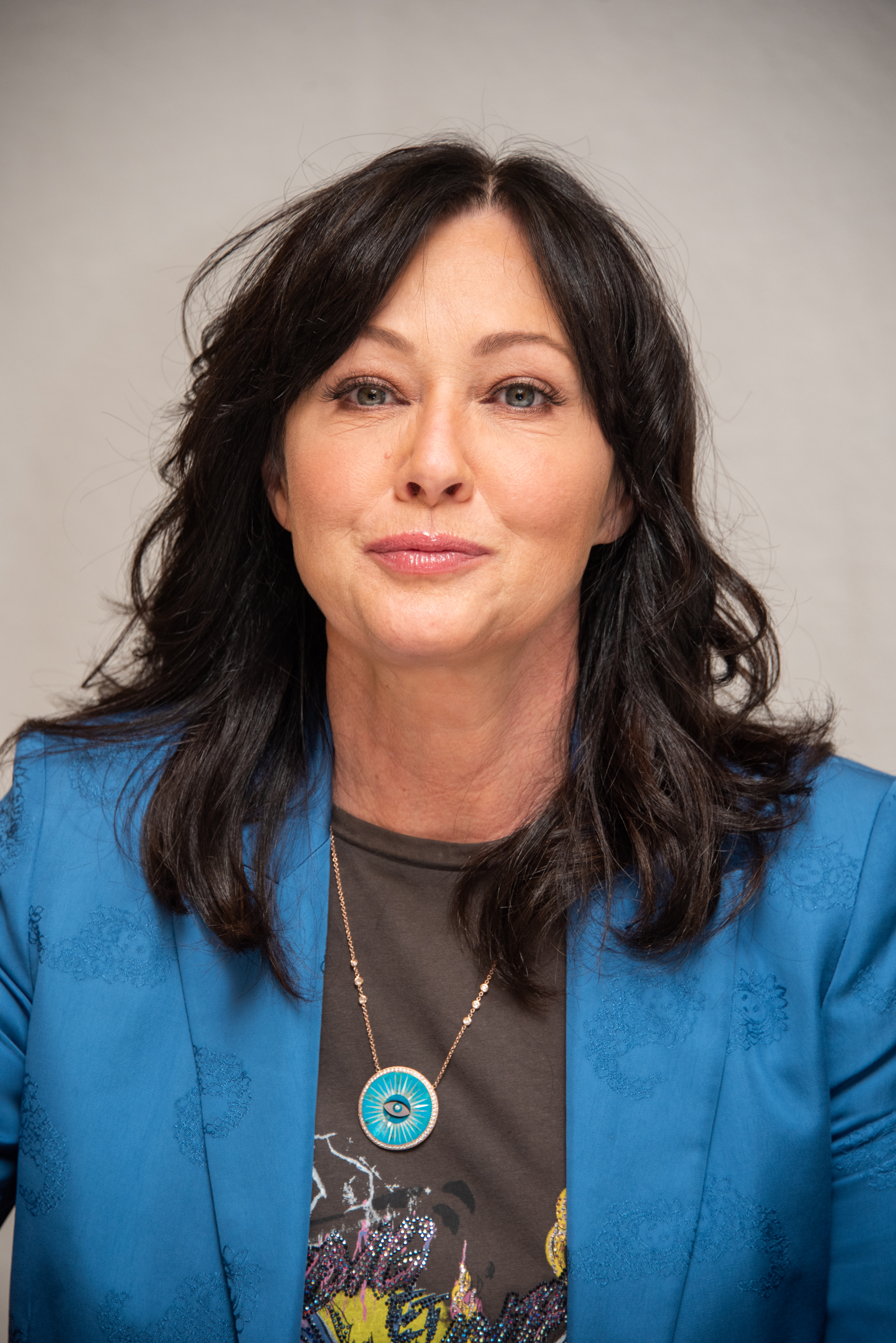 Shannen Doherty wearing a blue blazer over a graphic tee and a pendant necklace with a blue gem, smiling slightly at a press event