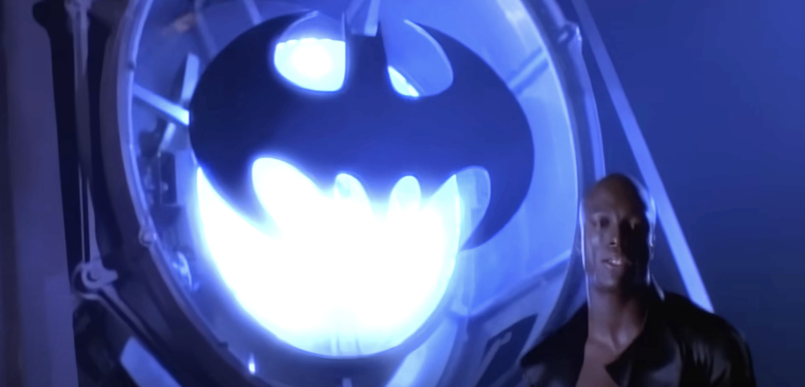 Singer Seal stands next to the Bat-Signal from Batman while serving a dramatic look, matching the theme of his hit song "Kiss from a Rose" in the background