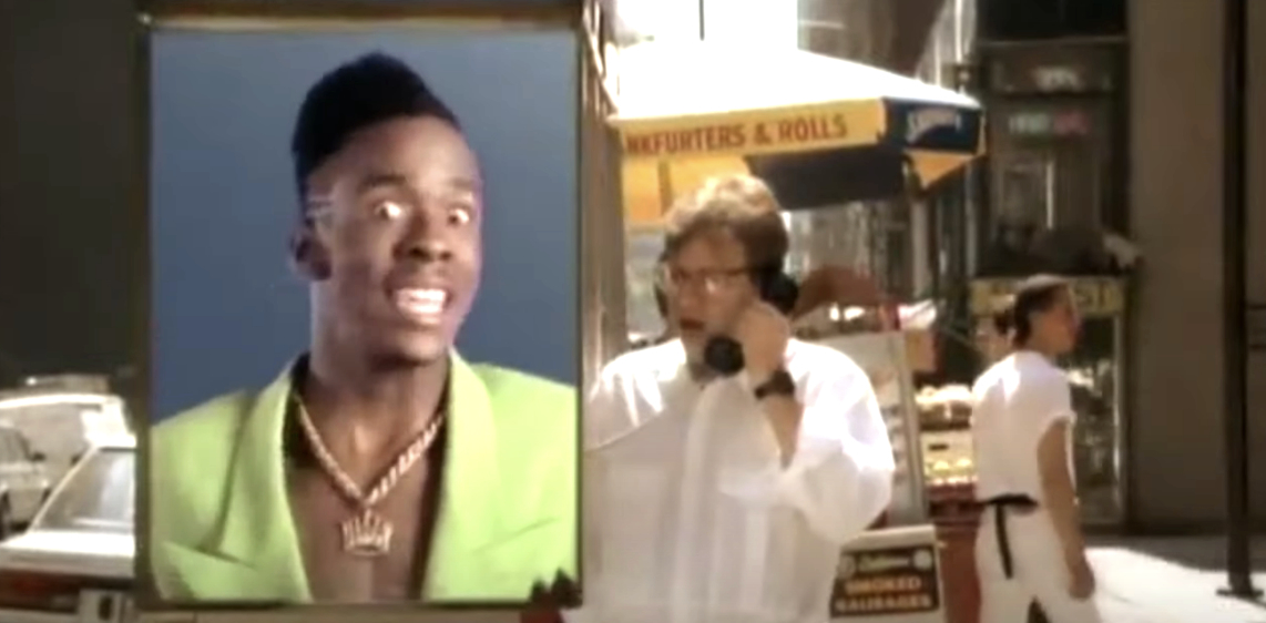 Man on left in large screen wearing a vibrant green suit and a gold necklace, man on right in white shirt speaking on a payphone. Urban street background