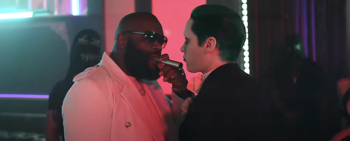 Rick Ross wearing a white suit and sunglasses is being interviewed by a man in a black suit holding a microphone