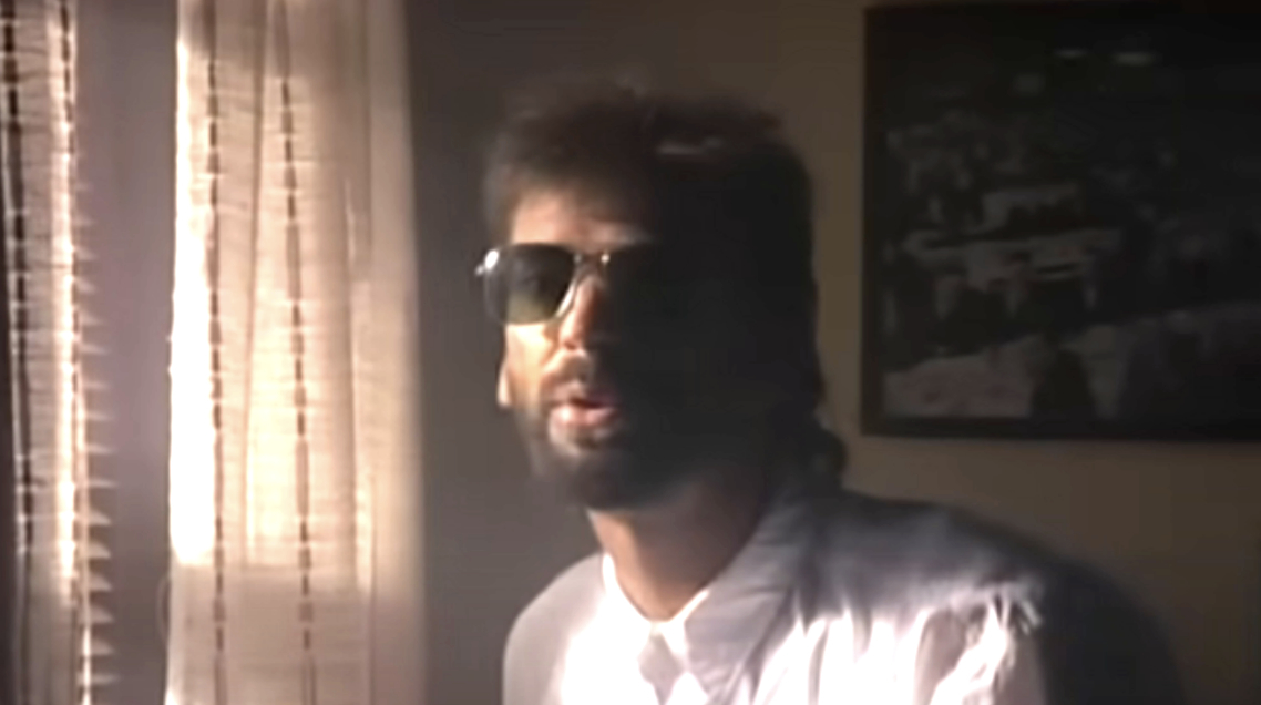 A man with sunglasses and a beard stands indoors near a window with blinds, wearing a white shirt. He exudes a cool and relaxed demeanor