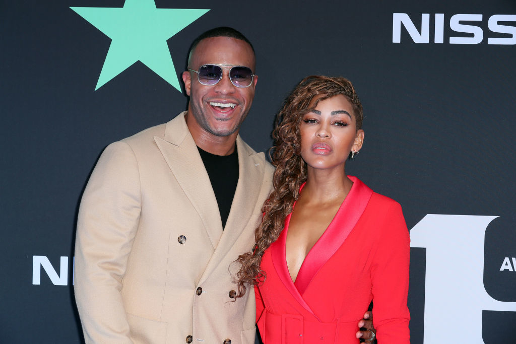 DeVon Franklin and Meagan Good at a red carpet event. DeVon wears a beige blazer, black shirt, and sunglasses. Meagan wears a deep V-neck red dress
