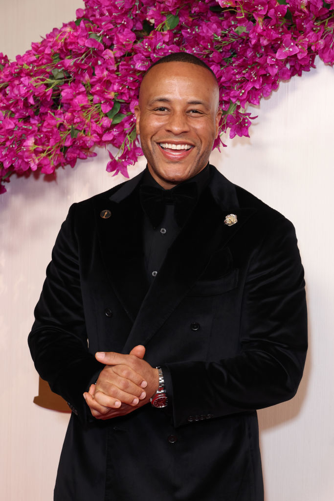 DeVon Franklin smiling in a velvet suit jacket, standing in front of a flower backdrop