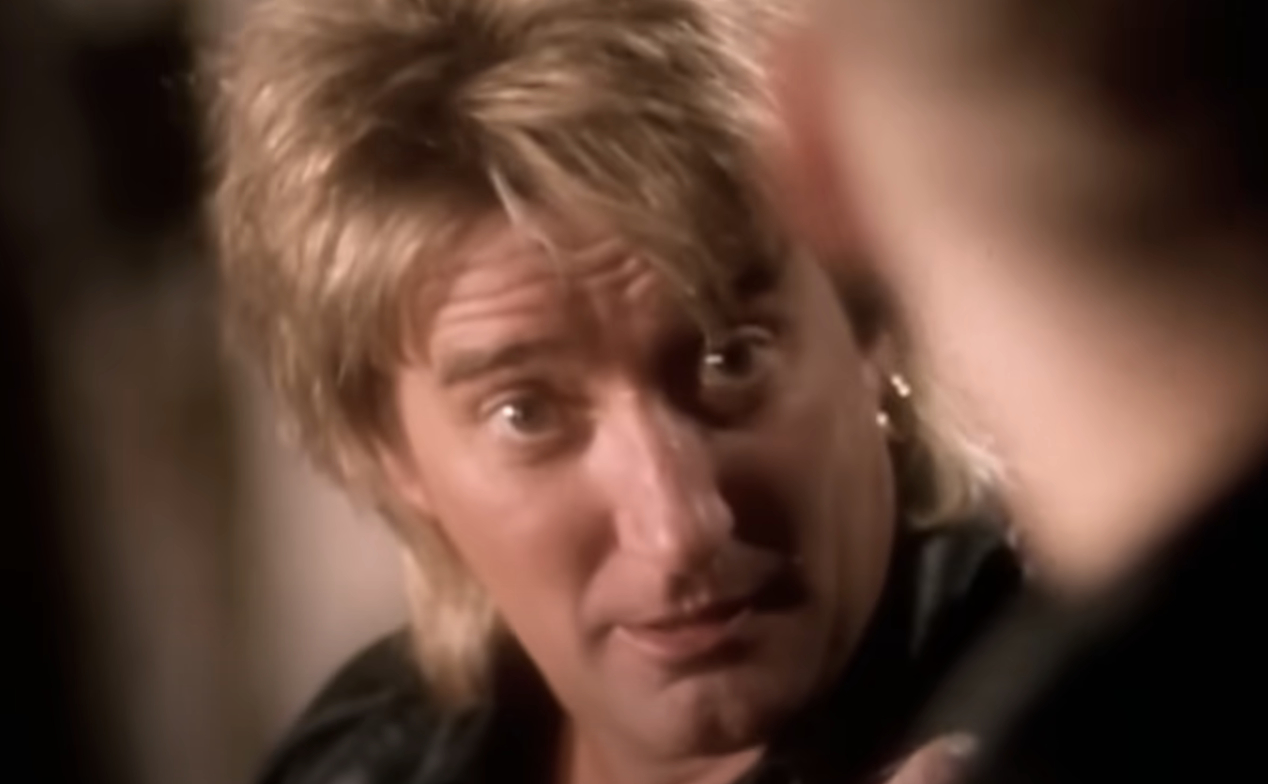 Rod Stewart, wearing a leather jacket, gazes attentively at someone off-camera in a music video scene