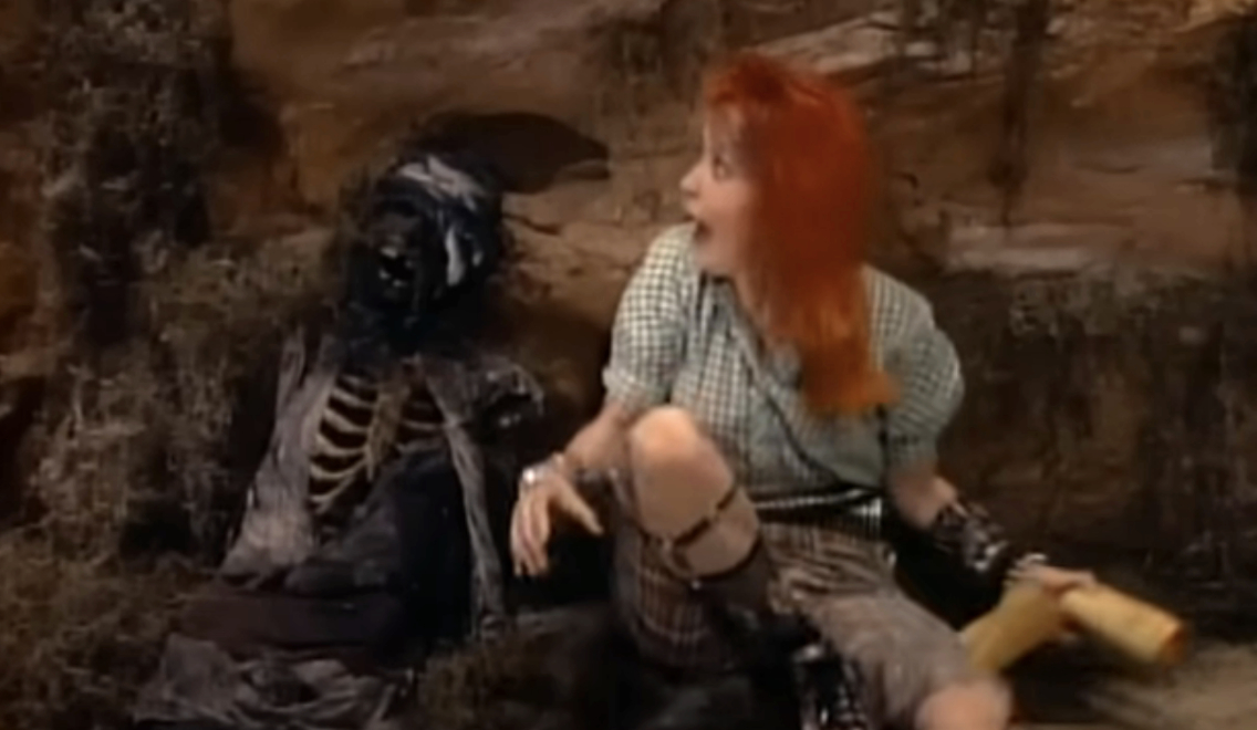 Cyndi Lauper, in casual checkered clothing, sits next to a skeleton on a set resembling a cave. She holds a scroll, appearing surprised or startled