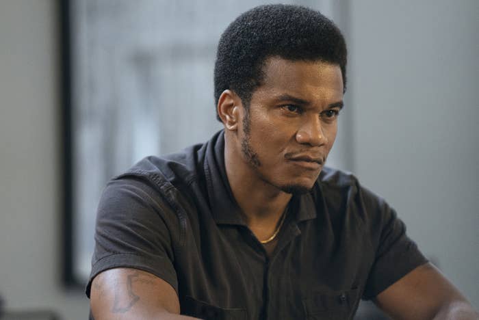Cory Hardrict sitting at a table with a serious expression wearing a short-sleeved button-up shirt