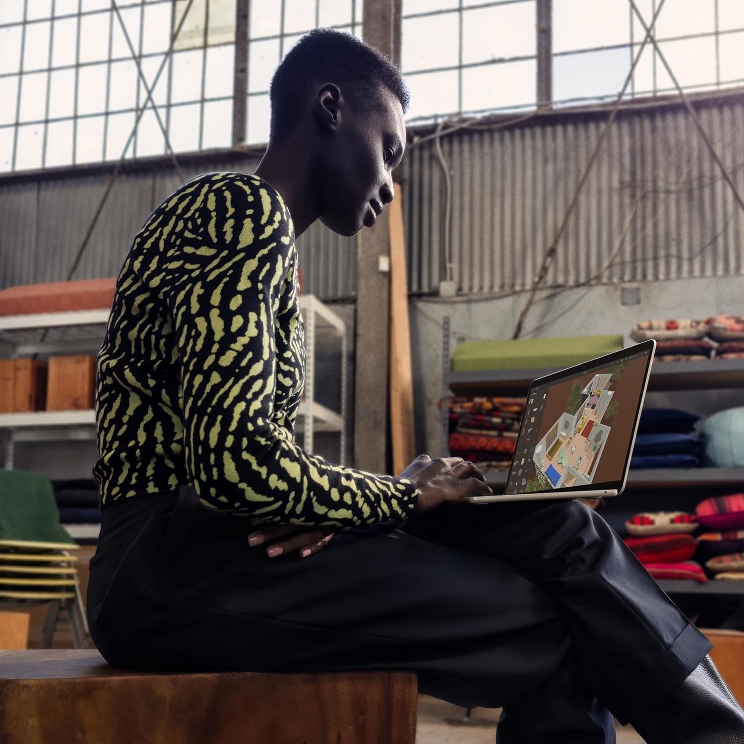 Person in a patterned top and dark pants uses a laptop in a modern, industrial-styled interior