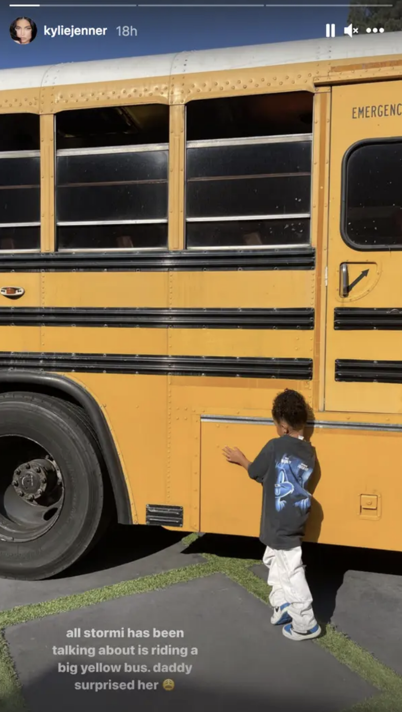 Stormi stands next to a large school bus. Caption by Kylie Jenner reads, "all Stormi has been talking about is riding a big yellow bus. daddy surprised her"