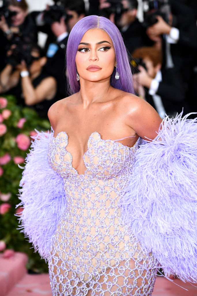 Kylie Jenner on a red carpet wearing a strapless, sheer gown adorned with intricate patterns and a dramatic feathered shawl, with long, straight hair