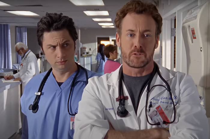 "Scrubs" characters JD and Dr. Cox are in a hospital hallway, both wearing medical scrubs and stethoscopes around their necks, with serious expressions