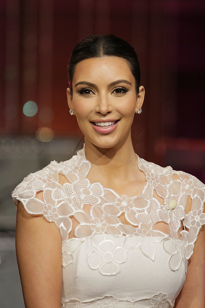 Kim Kardashian smiles, wearing an elegant lace dress with floral patterns. Her hair is pulled back in a sleek style, and she wears stud earrings