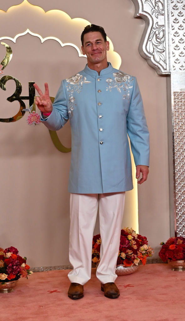 John Cena poses on the red carpet in a light, embroidered kurta and pants, giving a peace sign with his right hand